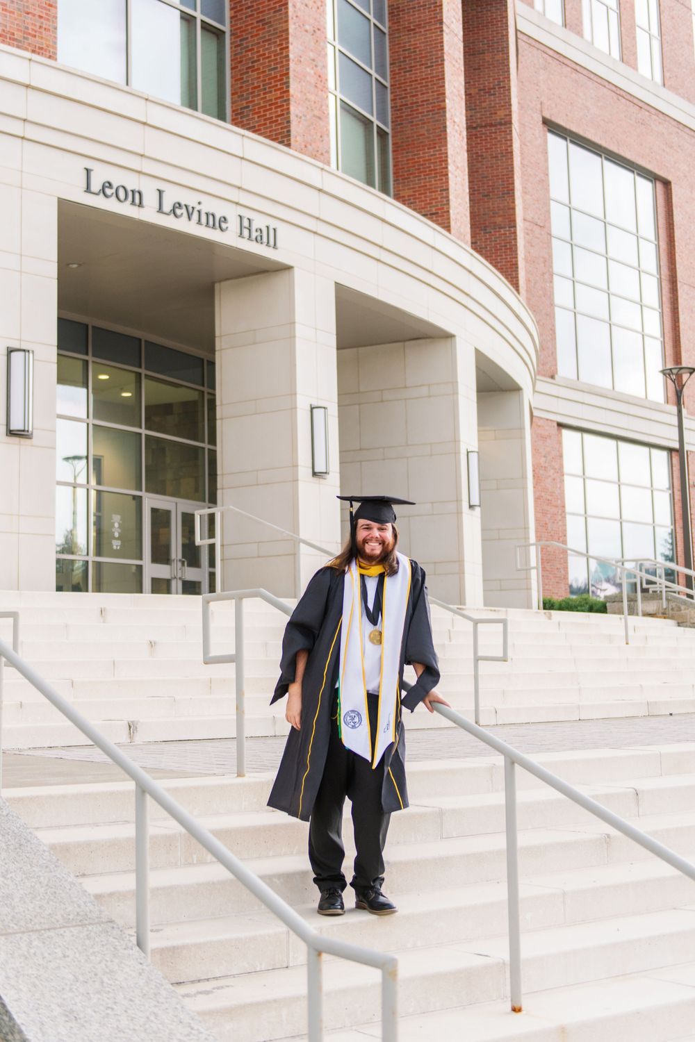 Graduate stands on steps of Leon Levine Hall wearing traditional graduation attire.