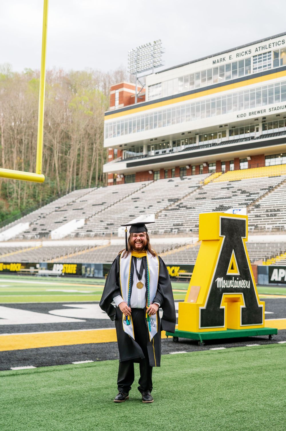 A graduate in full regalia stands proudly on a football field with stadium seating behind them.