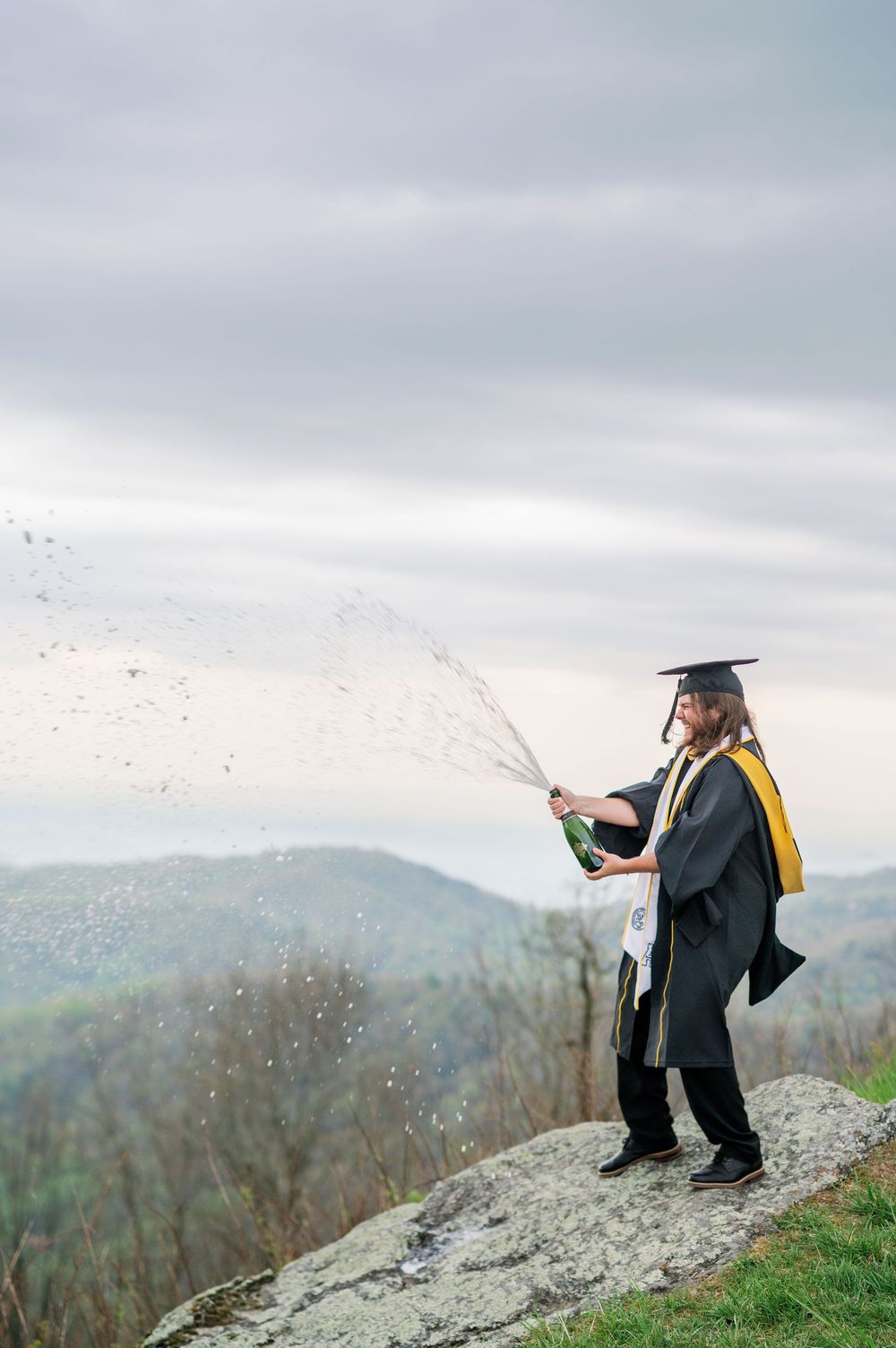 Graduate in cap and gown spraying champagne while standing on mountaintop overlook.