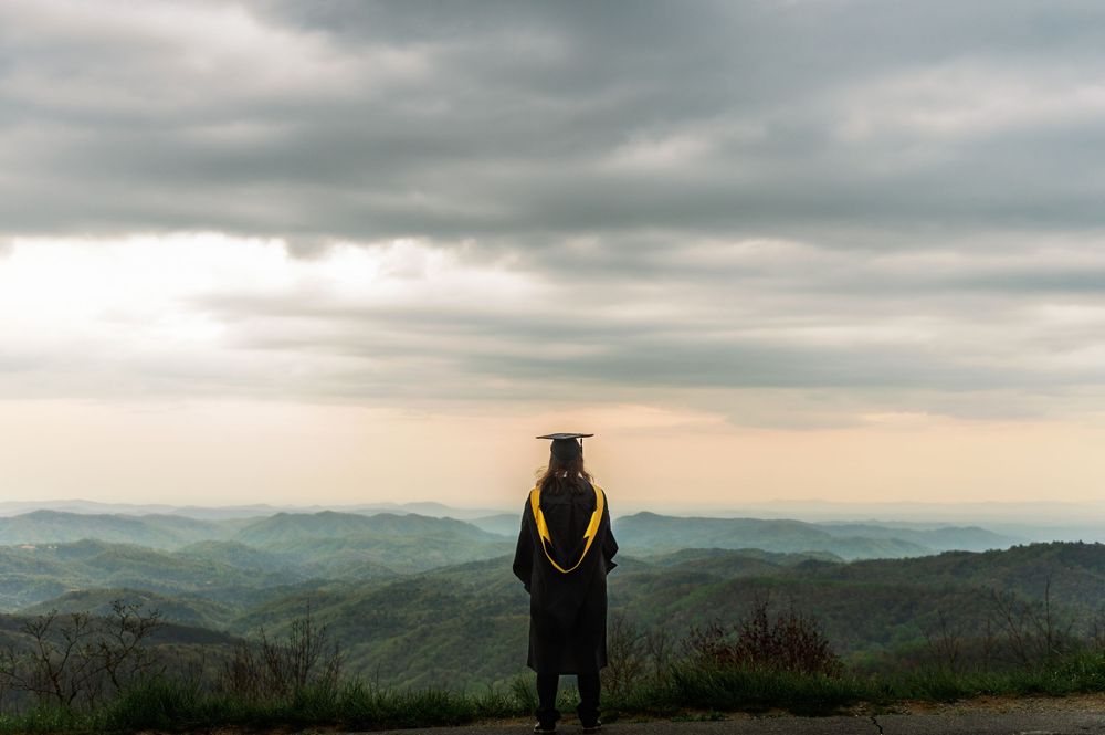 Graduate in academic regalia looks out over misty mountain landscape at sunset.