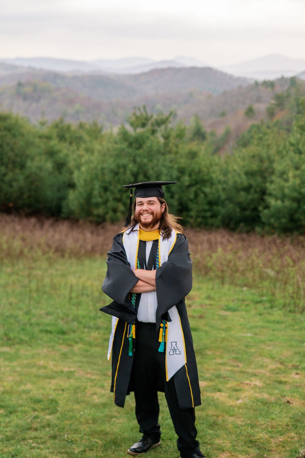 Graduate in academic regalia smiles while standing in mountain meadow.