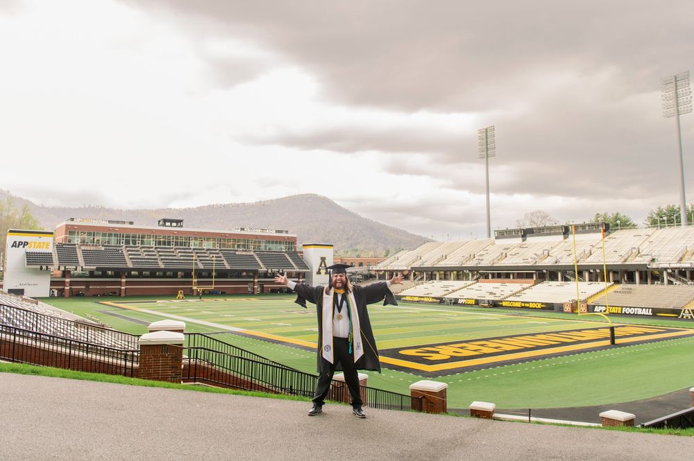 A graduate celebrates on an athletic field with mountains visible in the background.