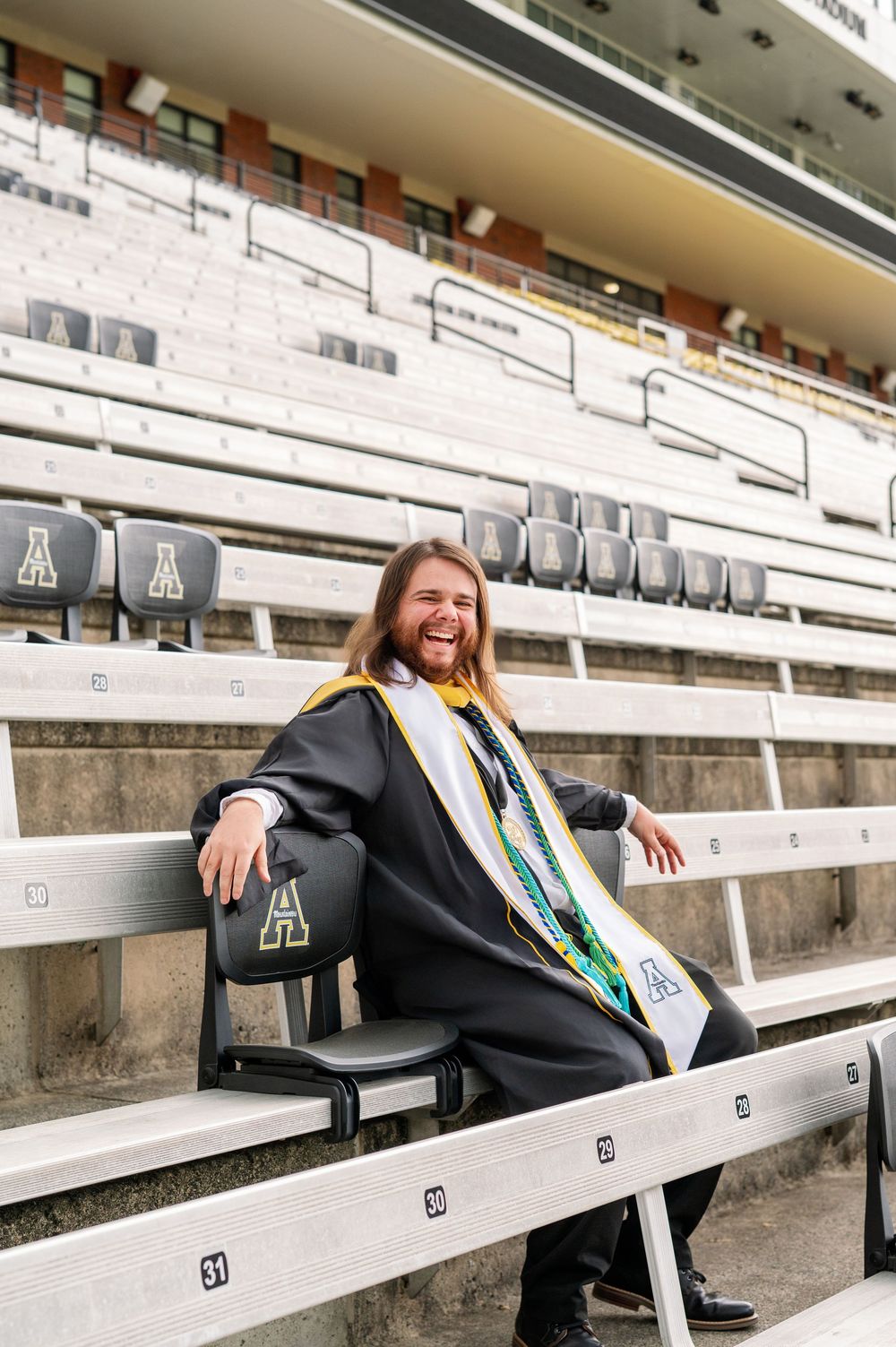 A graduate in academic regalia sits joyfully in stadium bleachers at Appalachian State.
