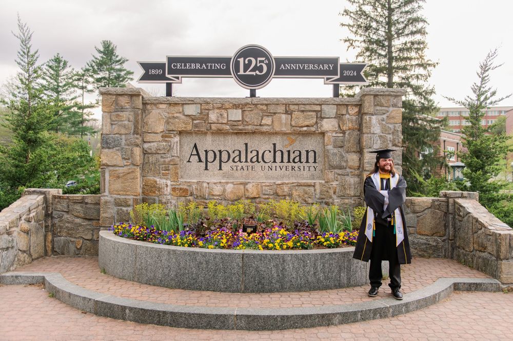 A graduate poses in front of the Appalachian State University 125th anniversary sign.