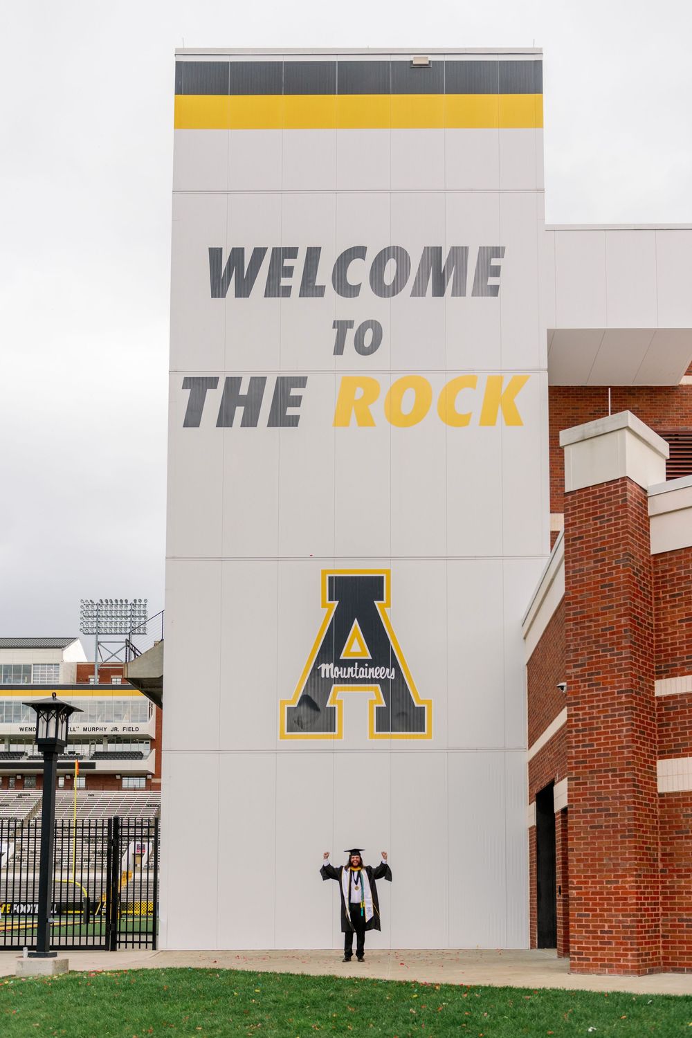 A graduate poses beneath a 'Welcome to The Rock' sign with Appalachian State University logo.