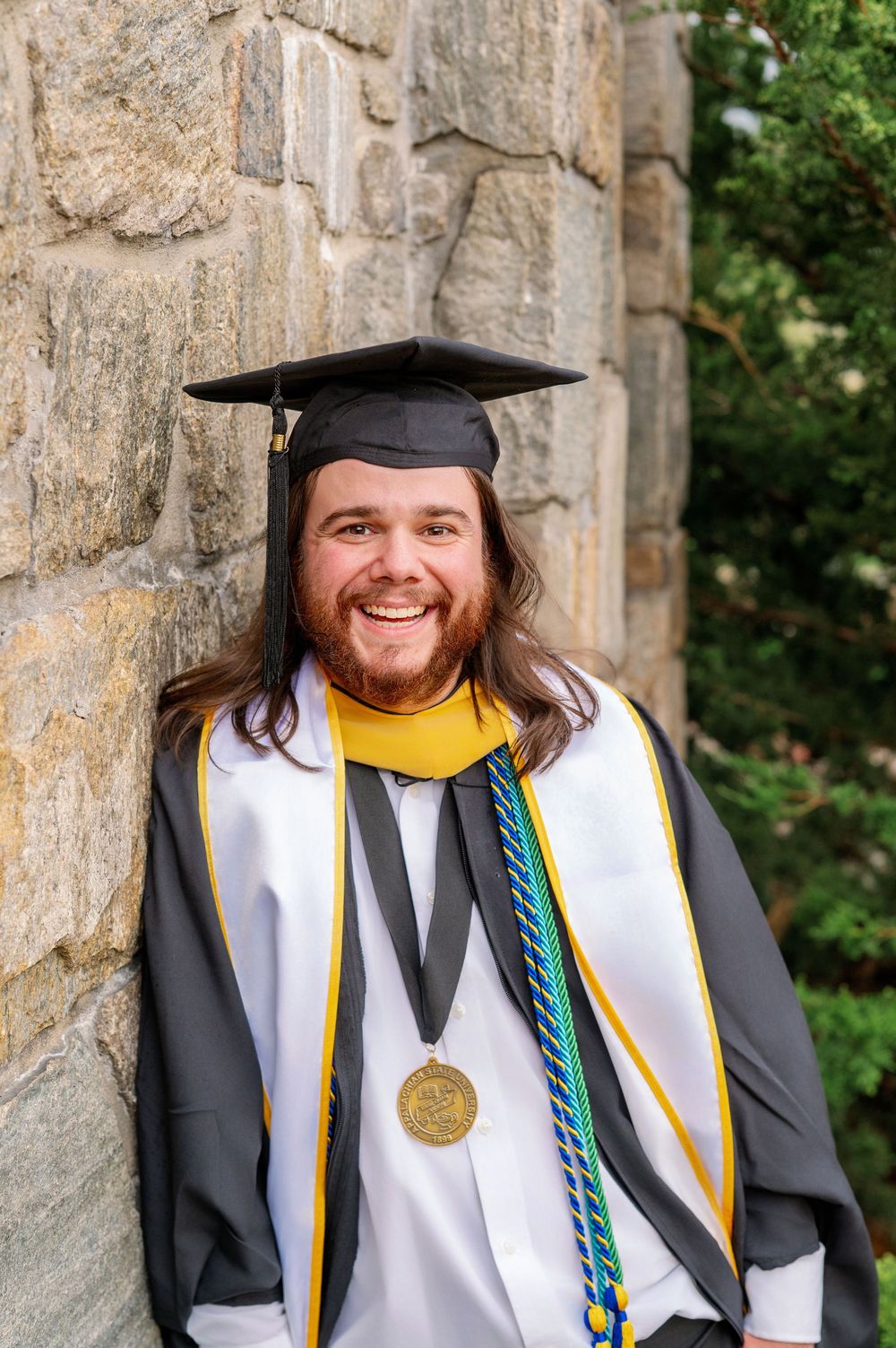 A graduate in academic regalia poses against a stone wall wearing honors cords and medals.