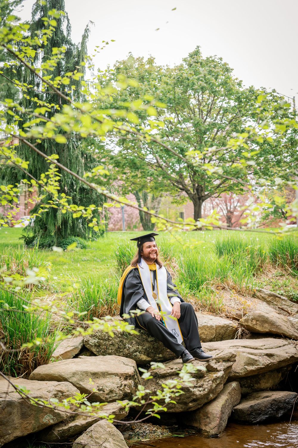 Graduate in cap and gown sits on rocky outcrop near stream in lush garden setting.