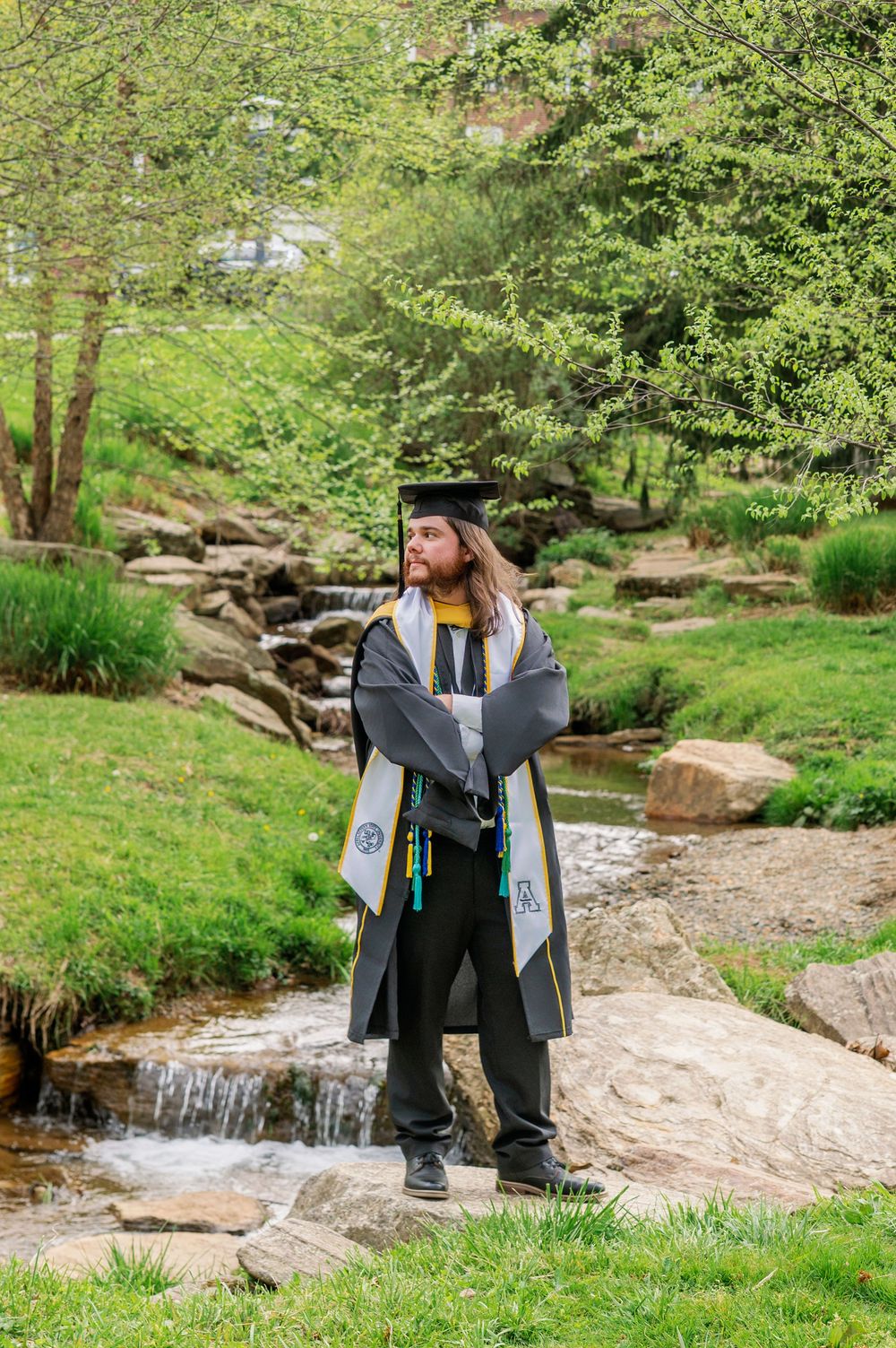Graduate poses near small waterfall wearing academic regalia in scenic outdoor garden.