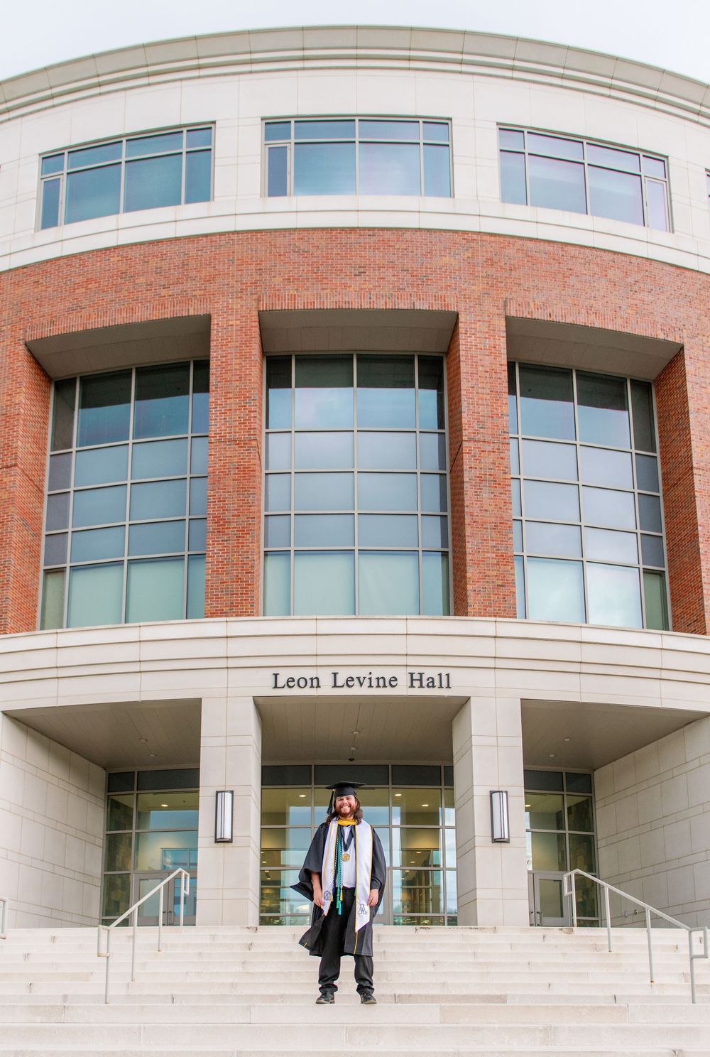 Graduate in academic dress stands in front of Leon Levine Hall brick building entrance.