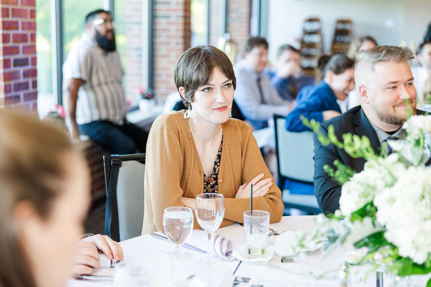 Dinner guests sit at an elegant table with white floral centerpieces in a bright restaurant setting.