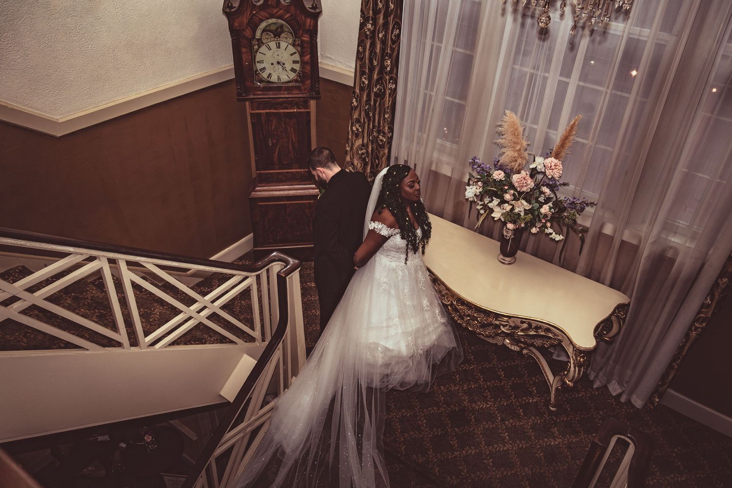 A person in a flowing white wedding gown descends an elegant staircase beside a vintage grandfather clock and ornate table.