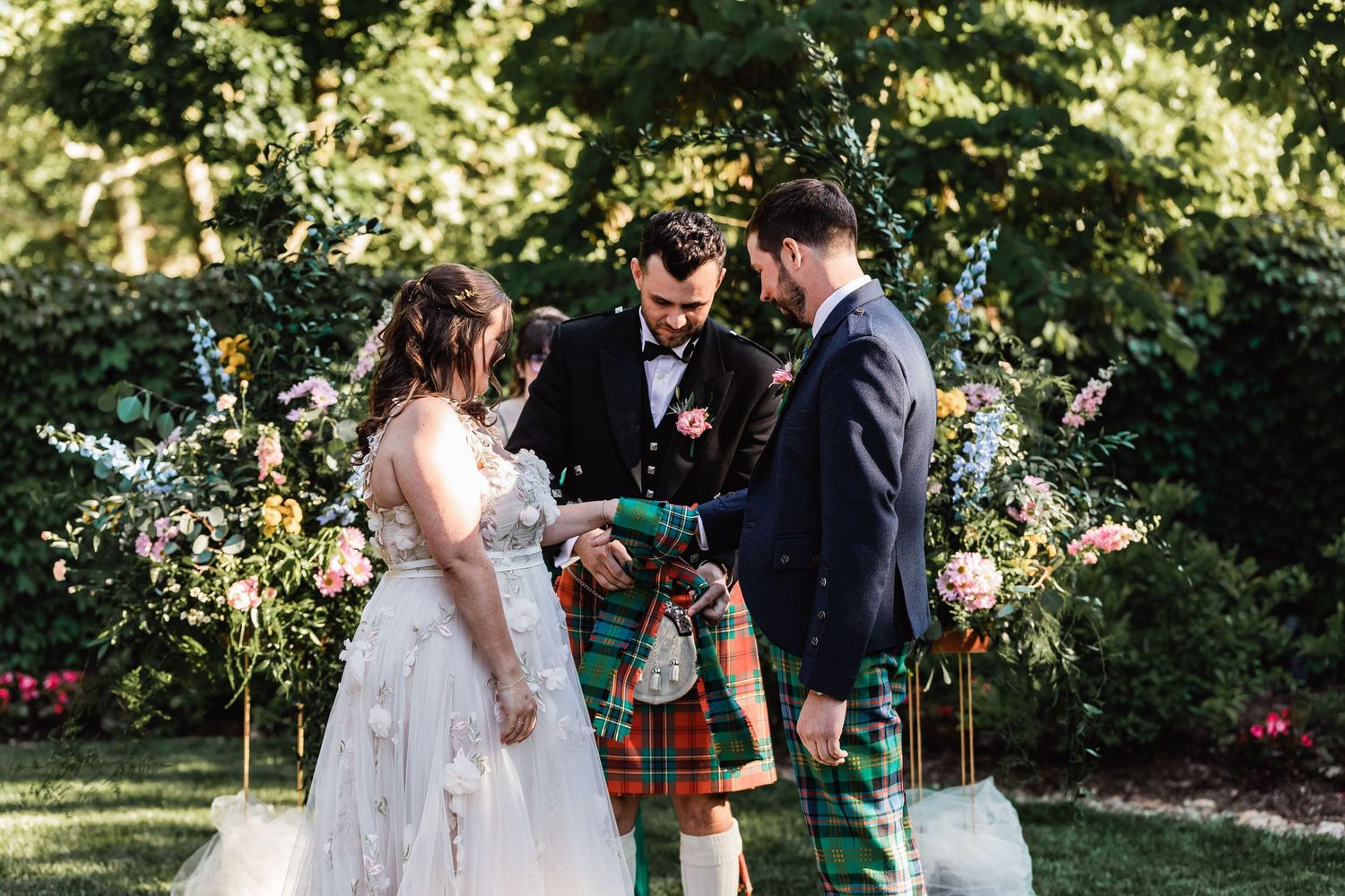 A wedding ceremony takes place outdoors in a garden with traditional Scottish kilts and formal attire.