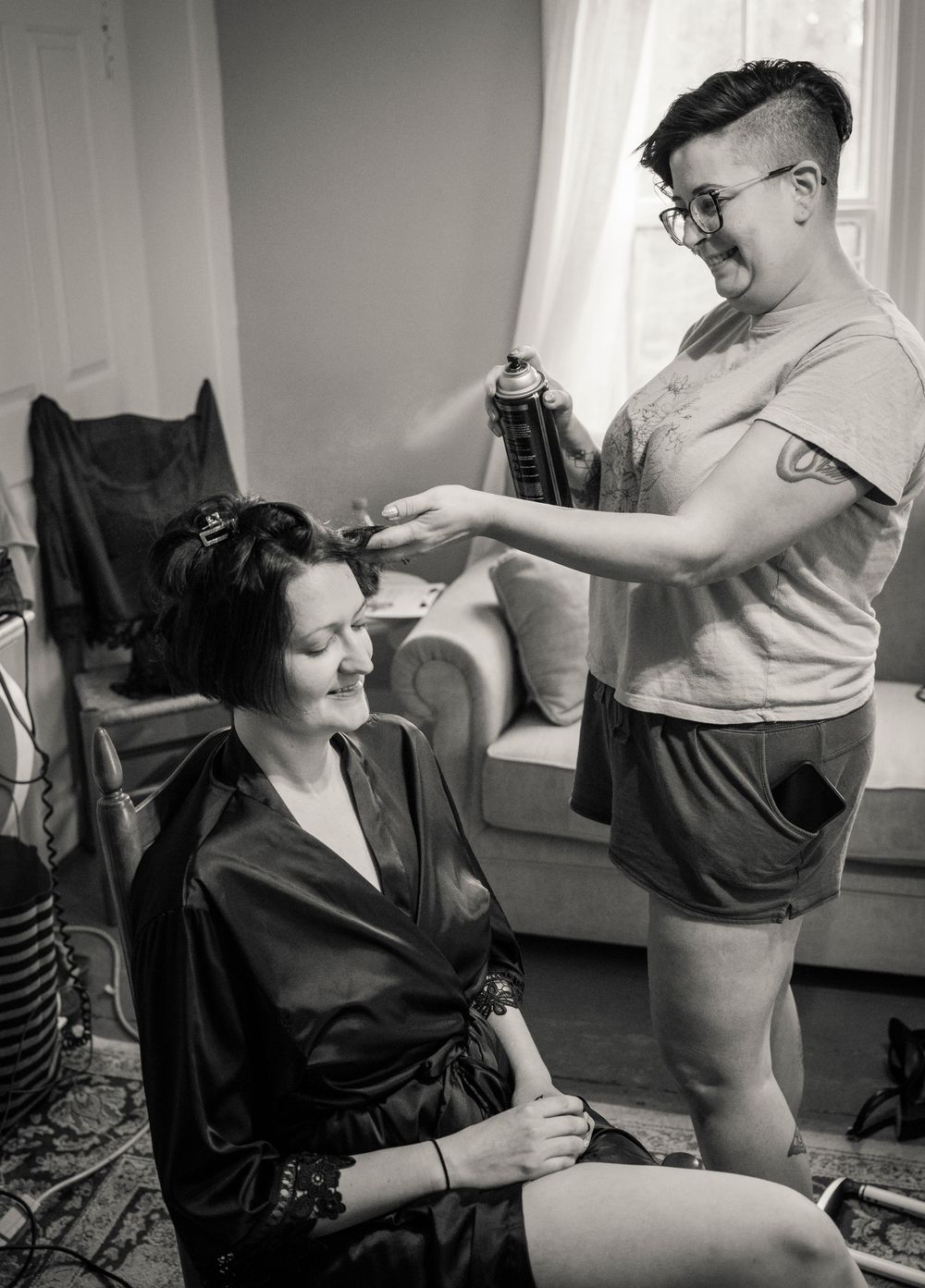 Vintage black and white photo of a hairdressing session showing a stylist in shorts and glasses styling a client's hair.