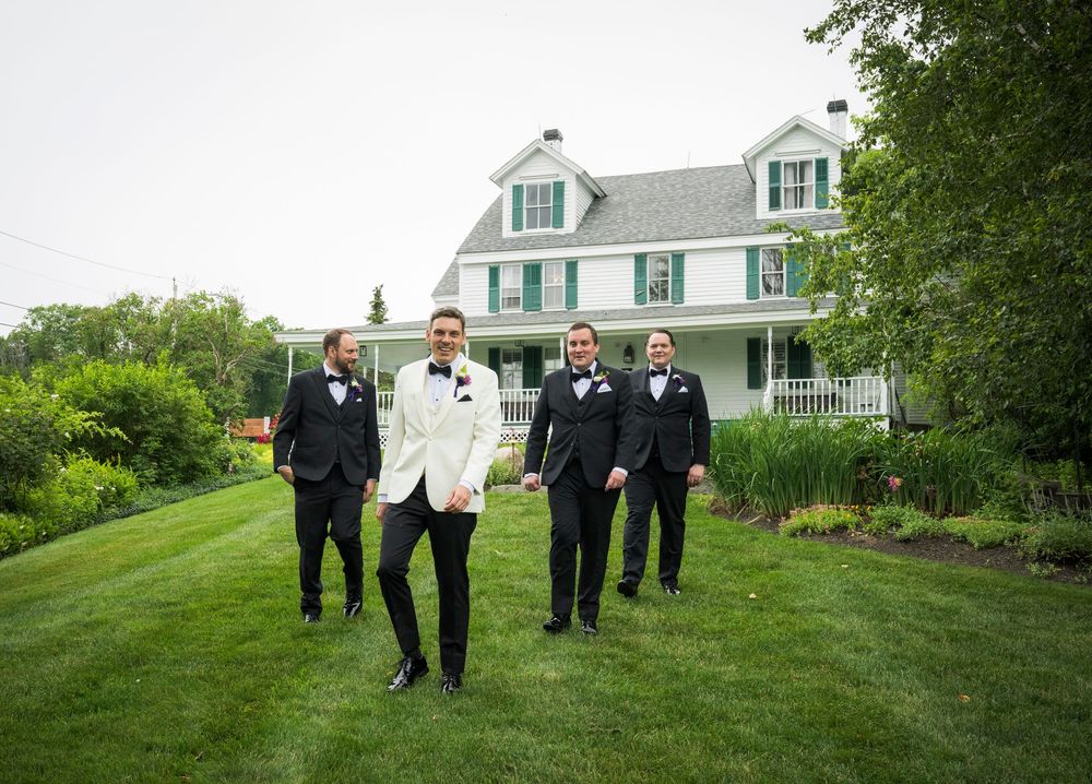 A wedding party in black suits walking on a grassy hillside with white houses in the background.