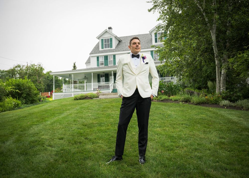 A person in a white tuxedo jacket stands on a manicured lawn in front of a grand Victorian-style house with wraparound porch.