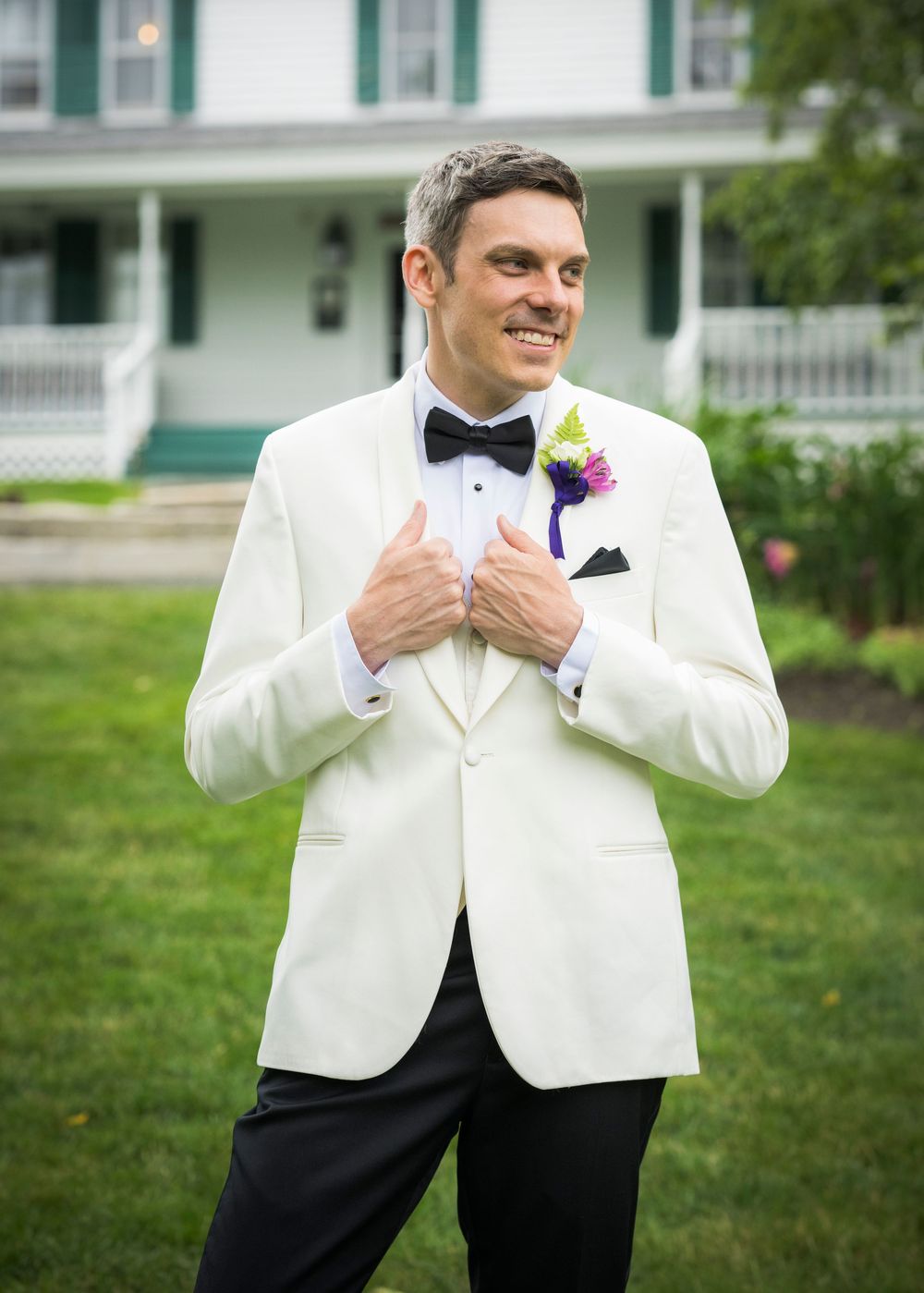 A groom in a formal white dinner jacket with black bow tie and purple boutonniere stands on a green lawn.