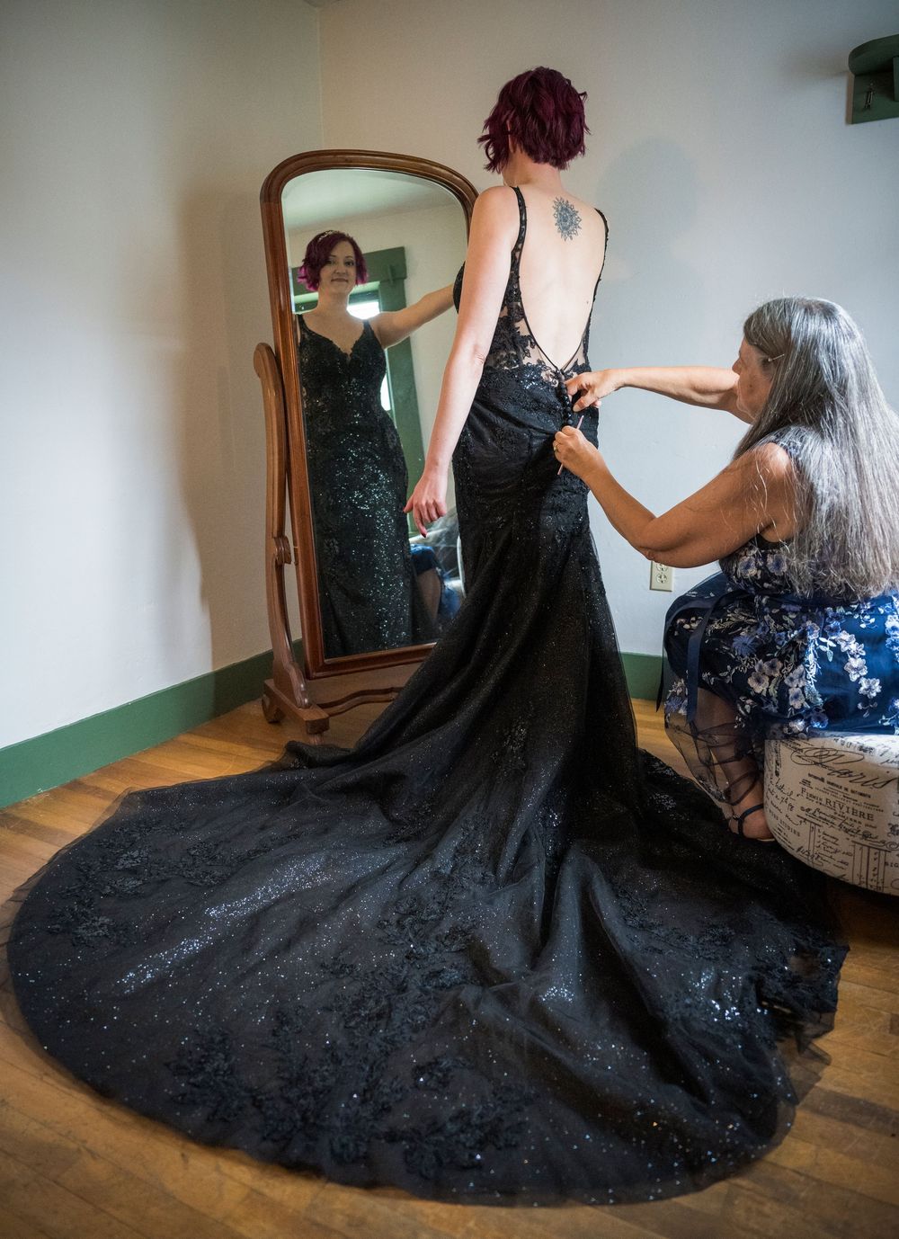 Someone tries on a stunning black lace formal gown with a dramatic train while getting fitted in front of a mirror.