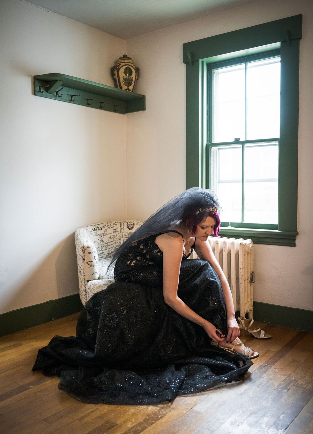 A person in an elegant black gown adjusts their shoe while seated in a minimalist room with natural light.