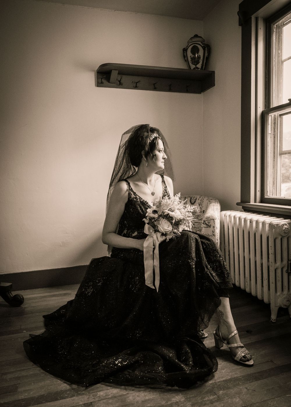 A figure in a black formal dress sits pensively by a window holding a bouquet of flowers in a moody monochrome scene.