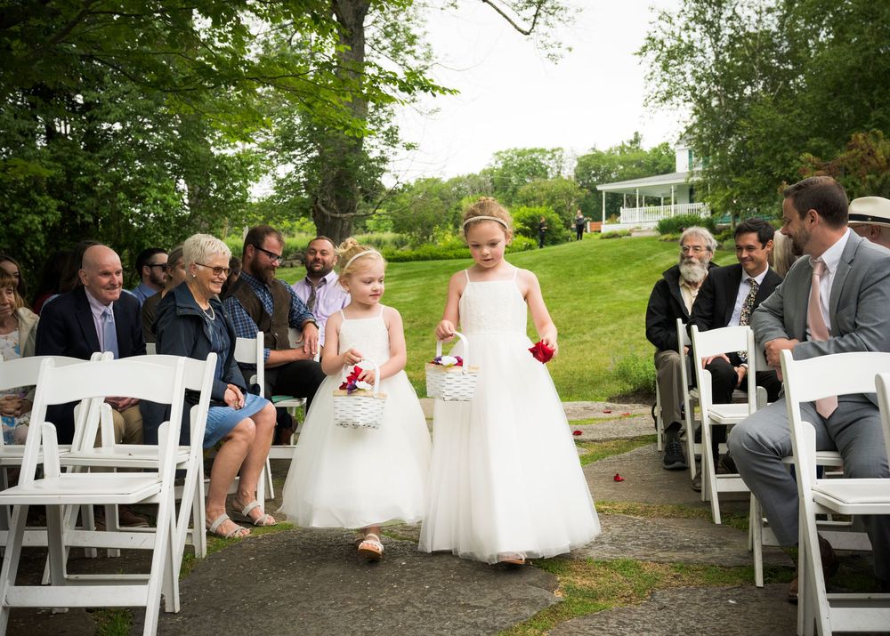 Two young children in white dresses walk down an outdoor path between rows of white chairs during a ceremony.