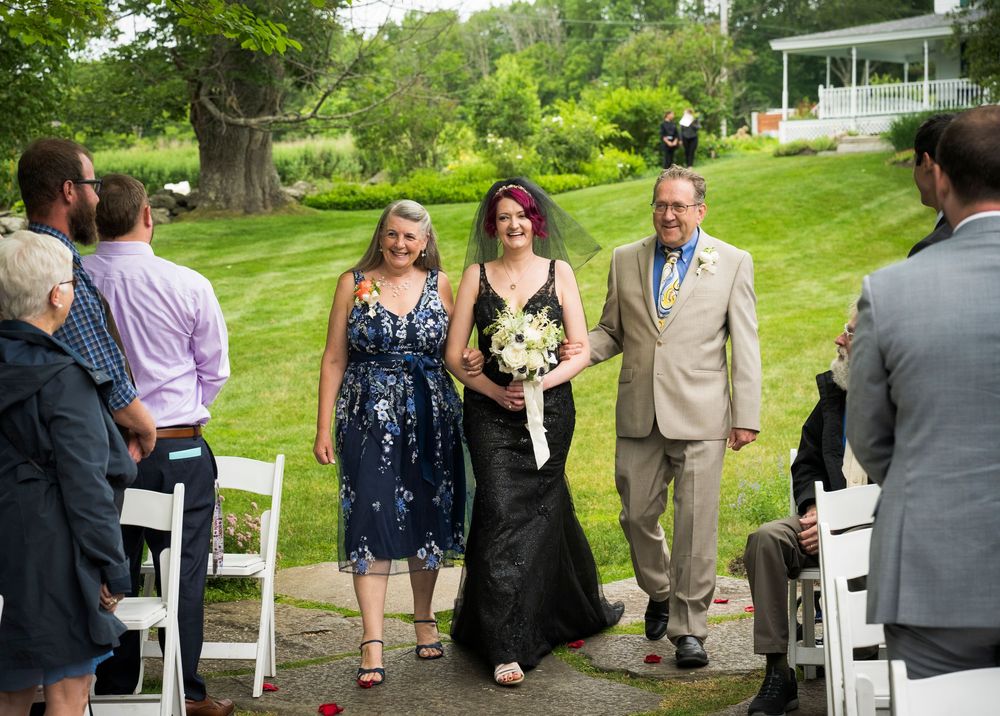 Wedding party members walking up the outdoor ceremony aisle on a sunny day with white chairs and green lawn.