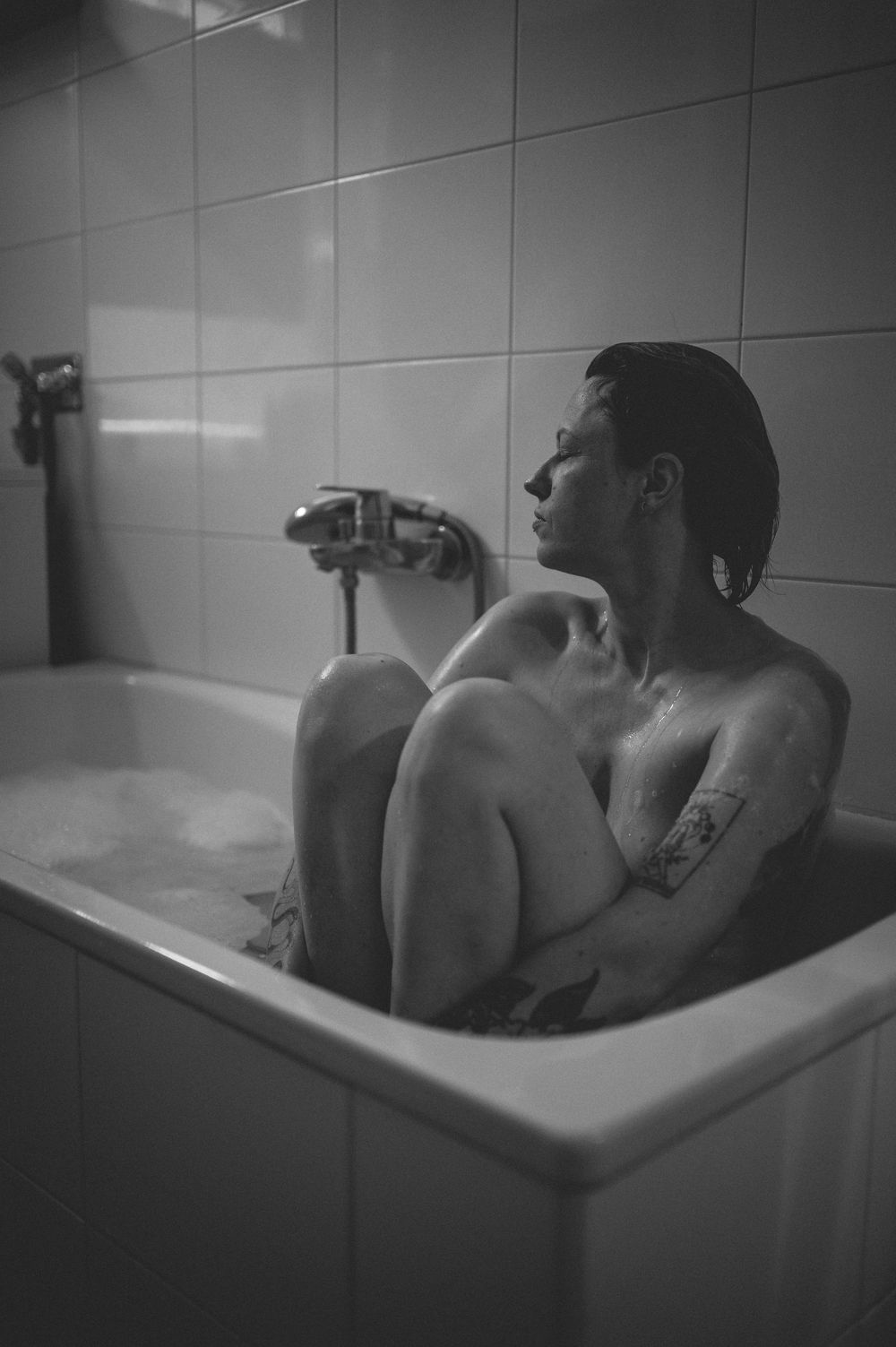 A person soaks in a white bathtub in a monochrome photograph, showing a tattoo on their body.