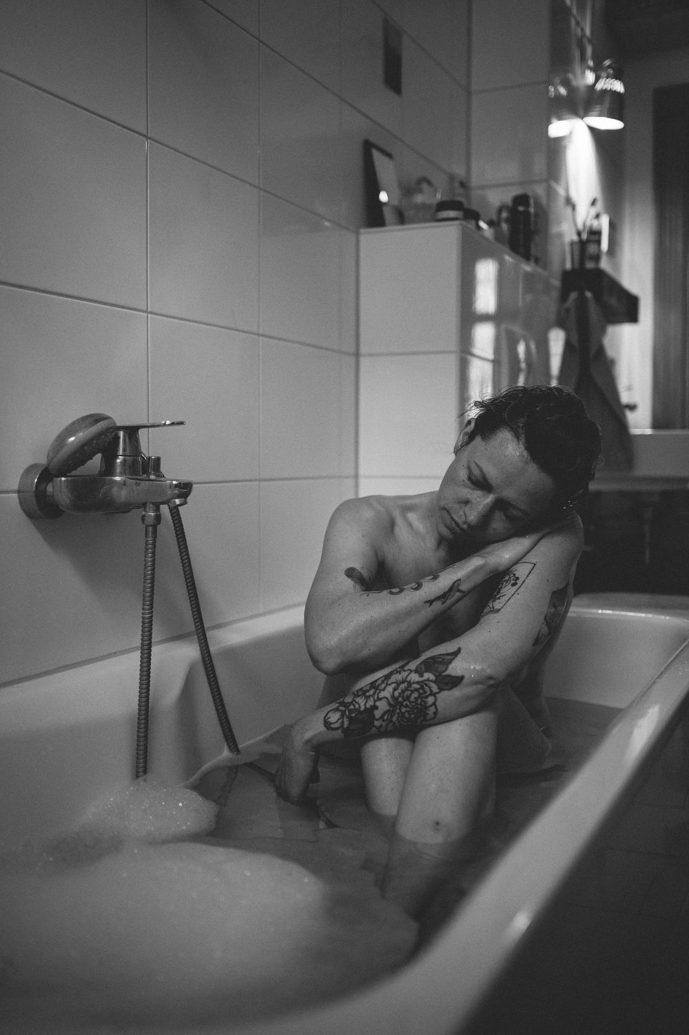 A person with tattoos relaxes in a bathtub in an artistic black and white photograph.