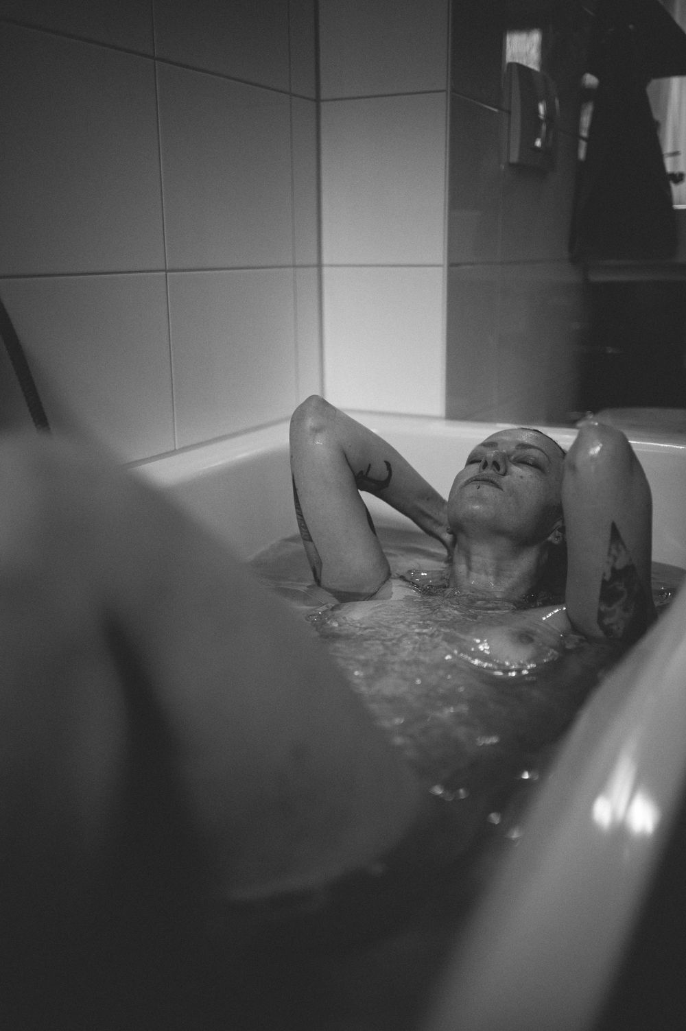 Black and white photo of someone relaxing in a bathtub with white tiled walls.