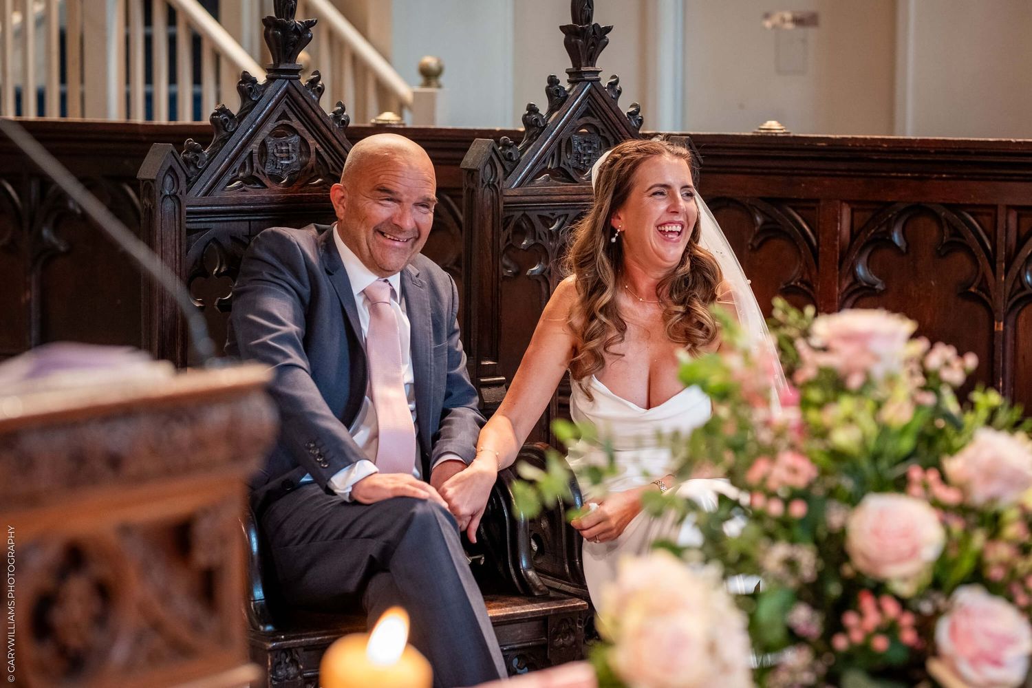Wedding couple laughs together while seated in ornate wooden church pew with pink floral arrangements nearby.