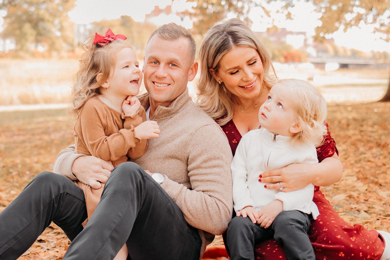 A cheerful family sits together outdoors on fallen autumn leaves during a warm sunset photoshoot.