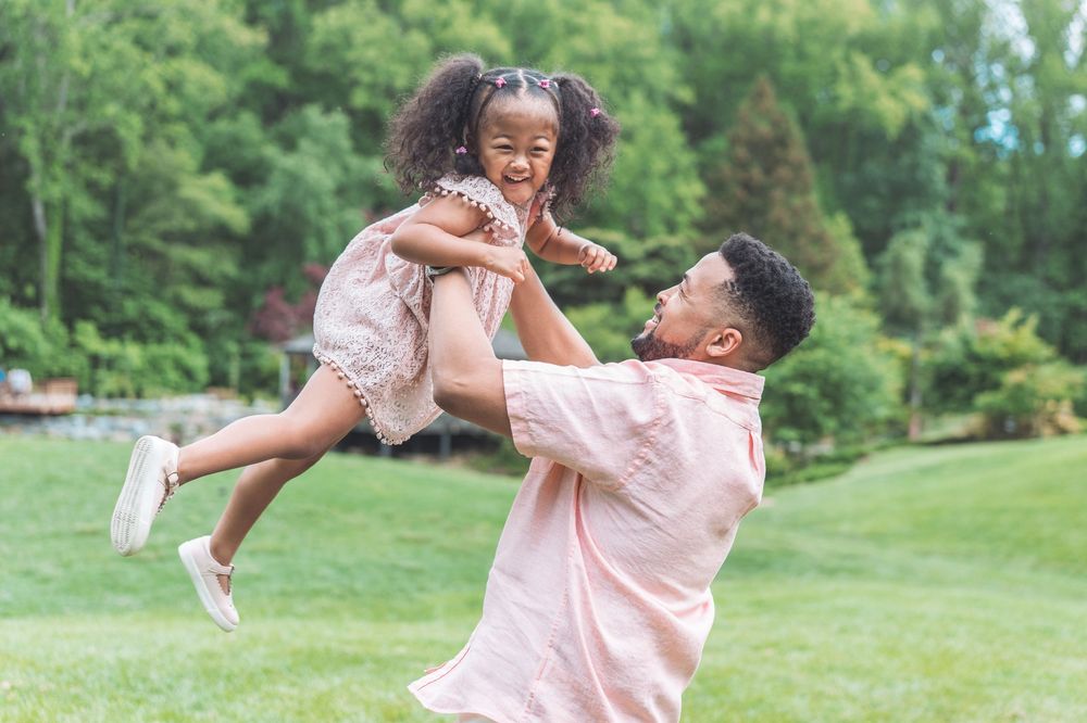 Parent playfully lifts and swings child in the air during outdoor playtime in a green park setting.