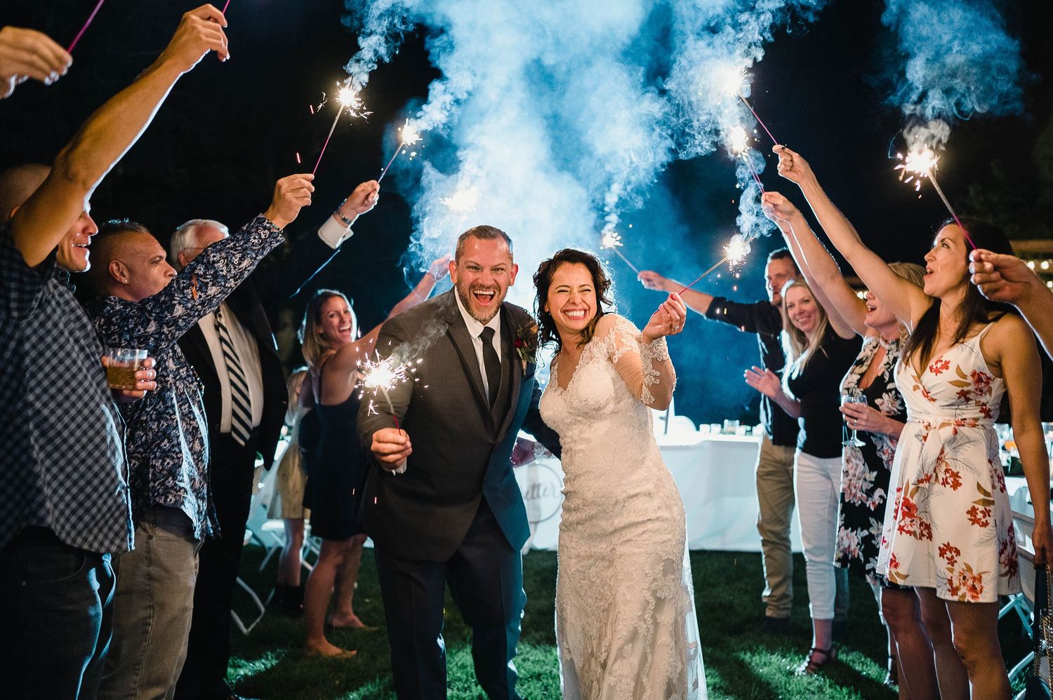 Wedding celebration at night with sparklers and blue smoke creating a magical atmosphere against the evening sky.