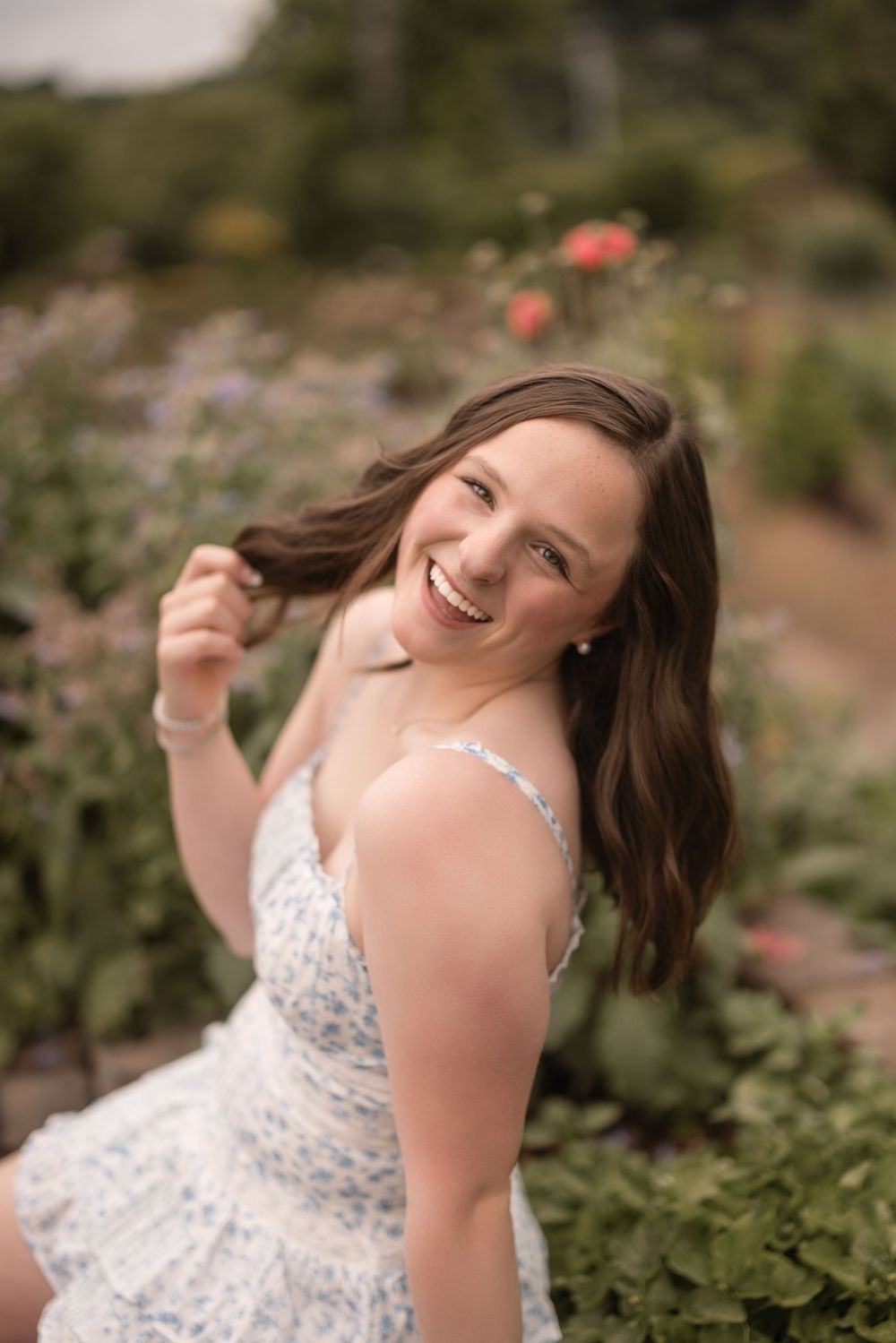 A person in a white lace dress smiles while sitting in a garden with blooming flowers in the background.
