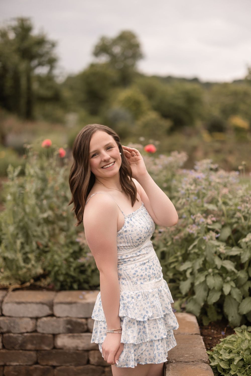 Someone in a white lace dress poses in a garden with stone walls and flowers on a summer evening.