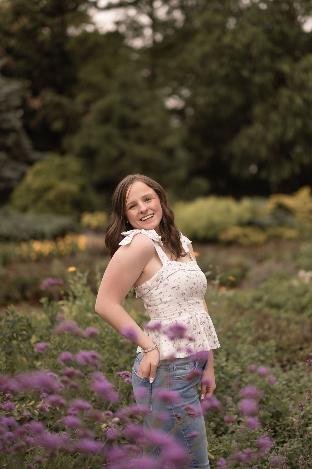 A person stands in a garden field with blooming purple flowers wearing a white lace top and jeans.