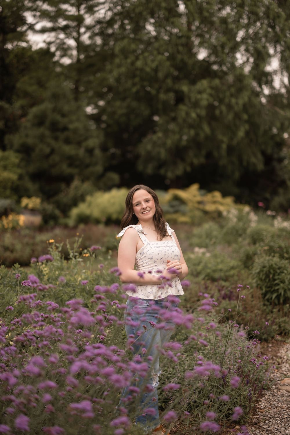 A white sleeveless top stands amidst a garden path with purple blooming flowers and lush greenery in the background.