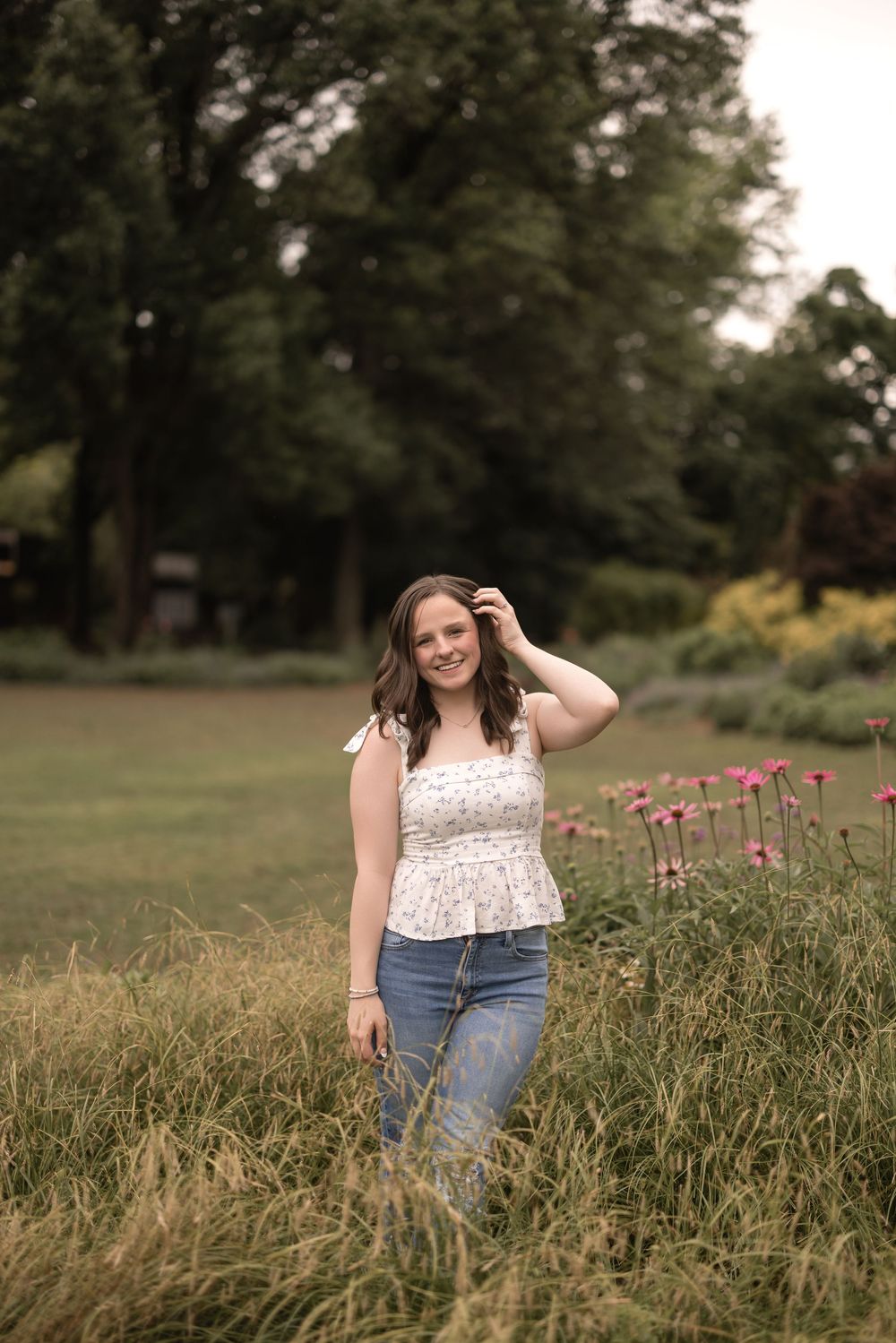 Someone stands in a lush green meadow wearing jeans and a white floral top as wildflowers bloom nearby.
