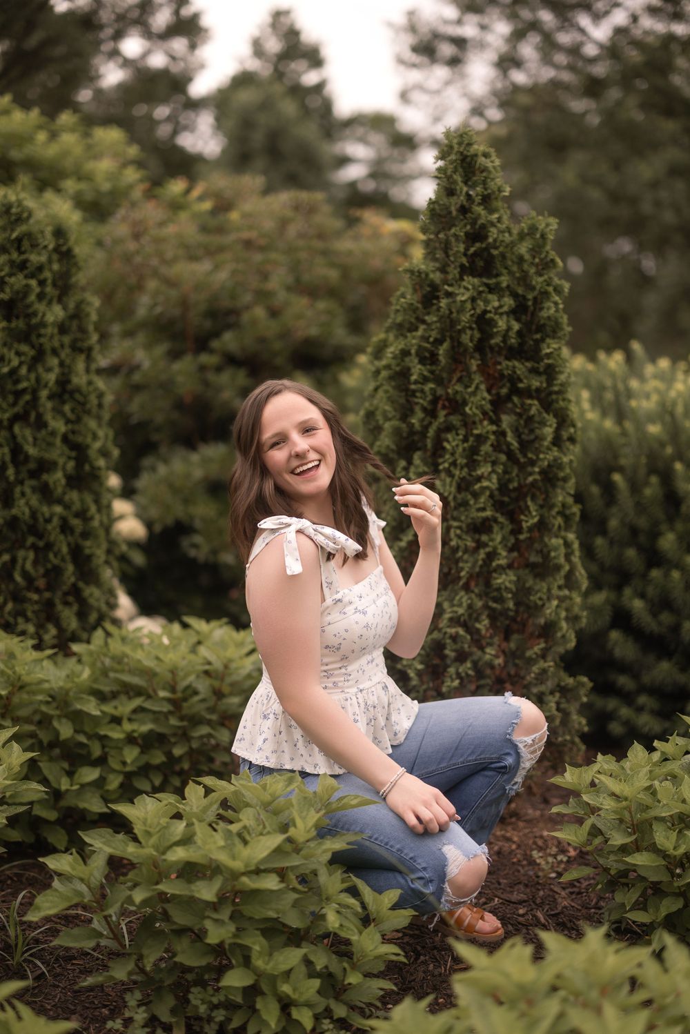 A joyful person in a white lace top and ripped jeans poses outdoors among evergreen shrubs and landscaping.