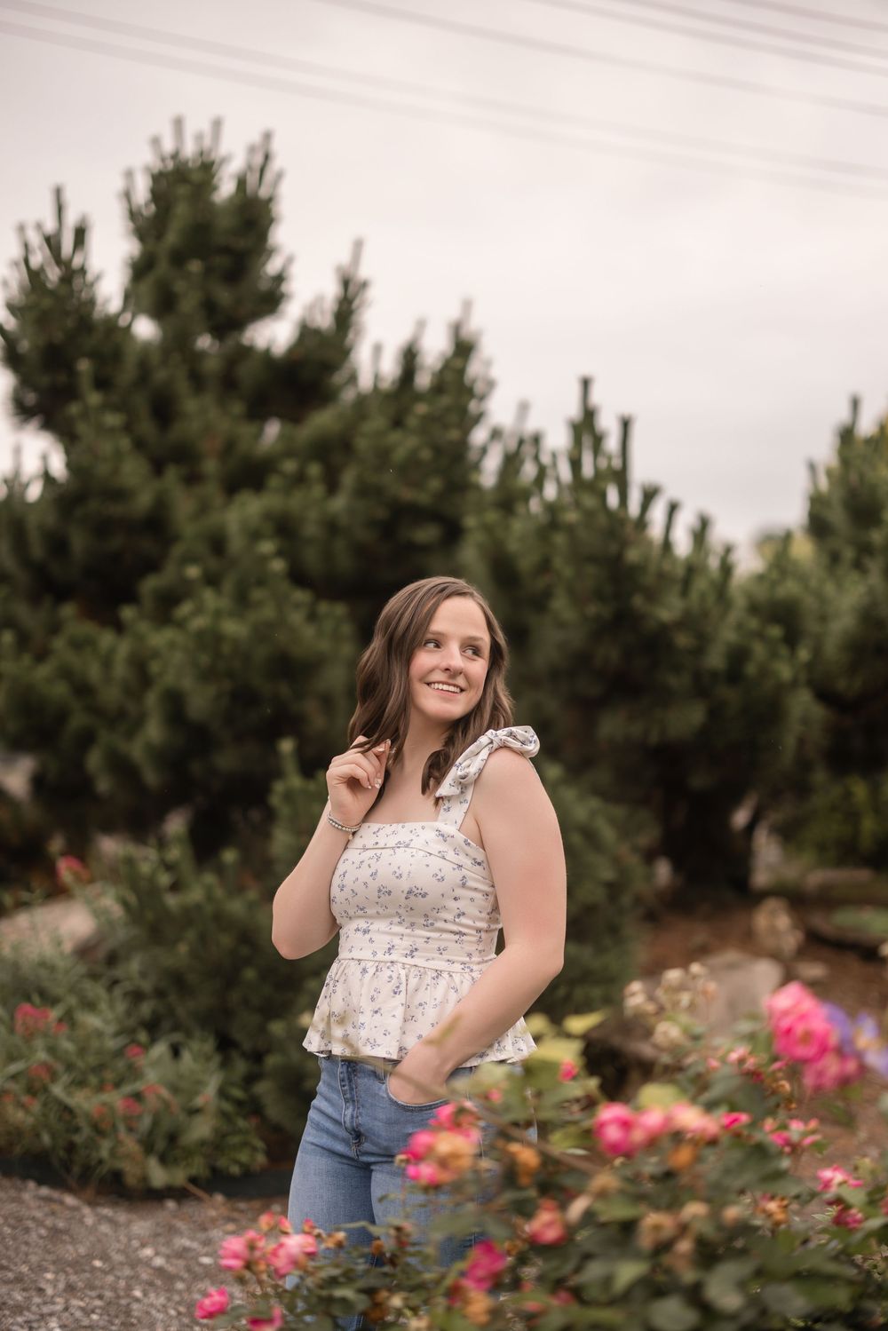 A woman in a white lace top and blue jeans stands in a garden with blooming pink roses and evergreen trees.