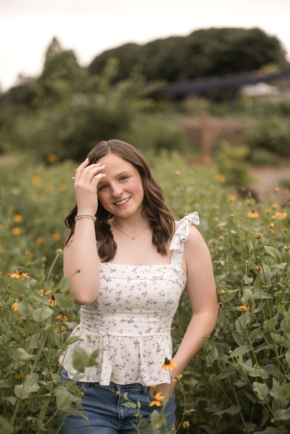 A person wearing a floral peplum top and jeans poses while smiling in a garden of wildflowers on a sunny day.
