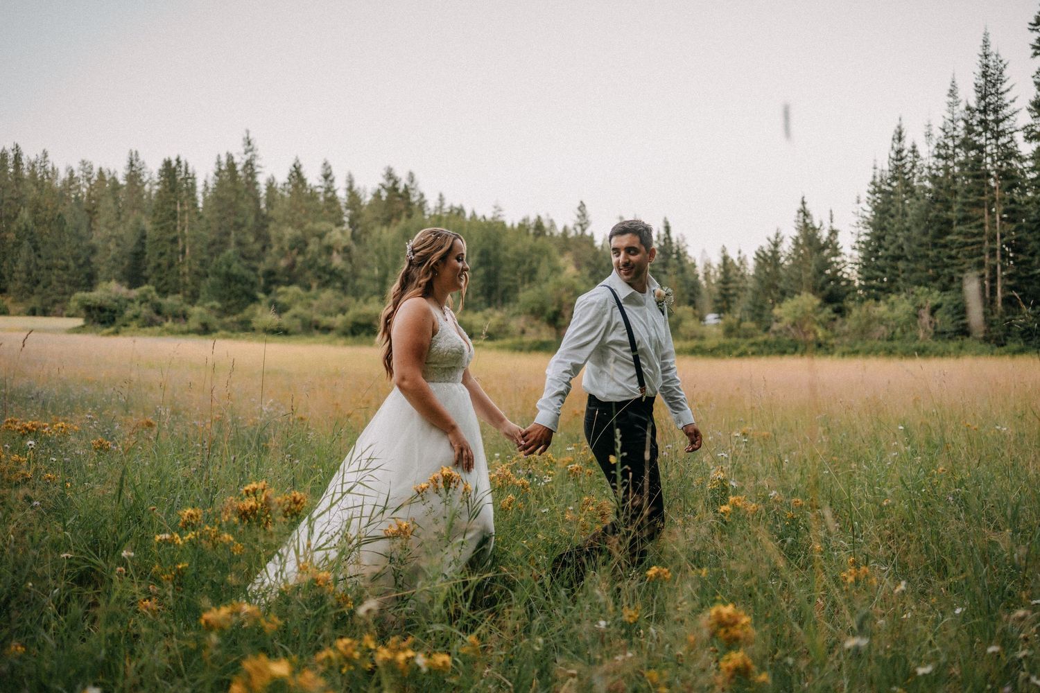 A couple walks hand in hand through a wildflower meadow surrounded by evergreen forest on their wedding day.