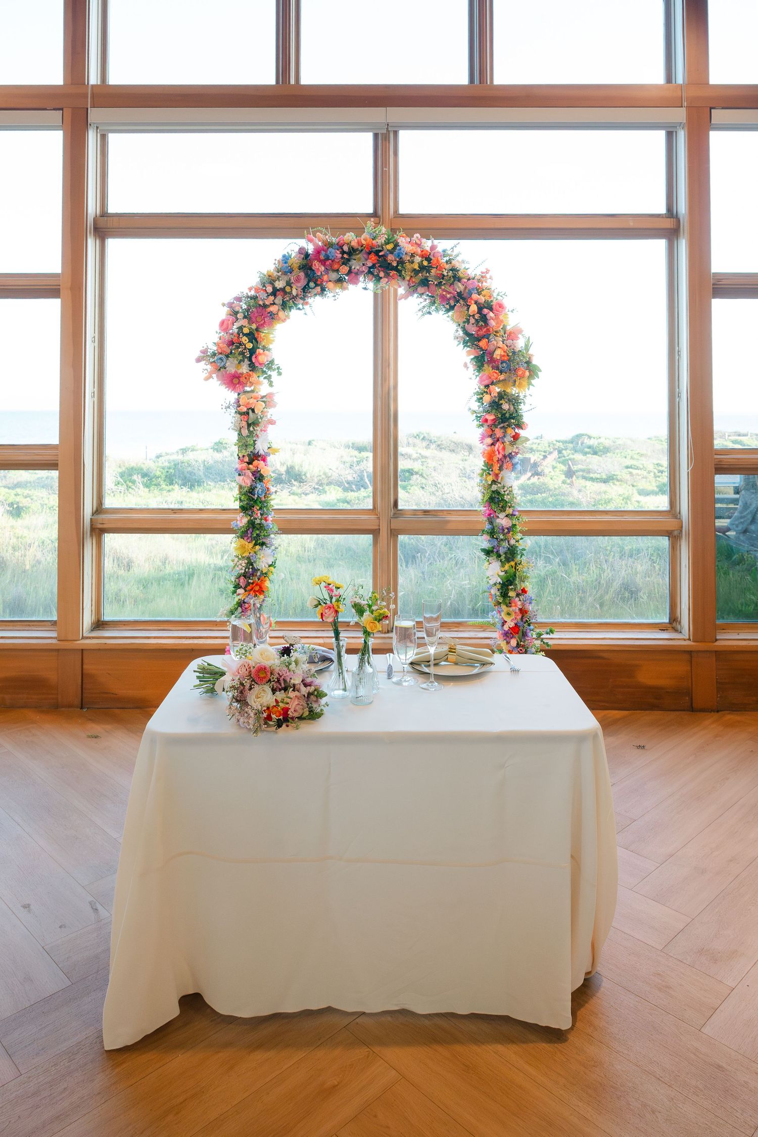 Floral arches line a row of white tables in a bright wooden room with large windows overlooking nature.