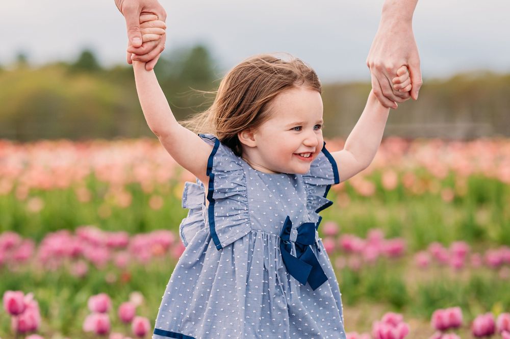 Happy child wearing blue dress holds hands with adults while standing in a vibrant pink tulip field on sunny day.