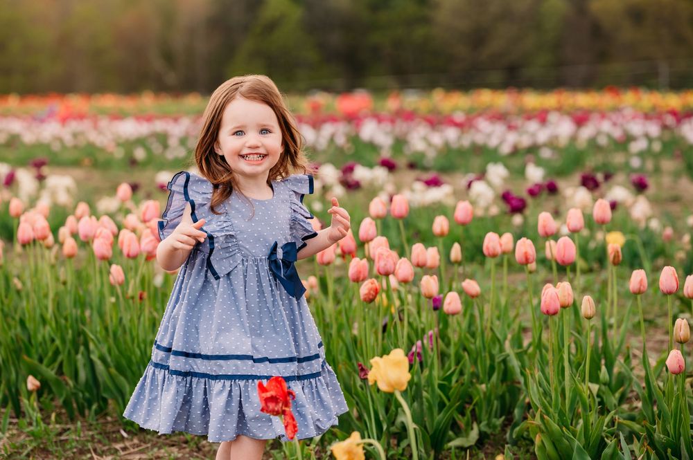 Young child in blue ruffled dress standing in colorful spring tulip field filled with pink and yellow flowers.