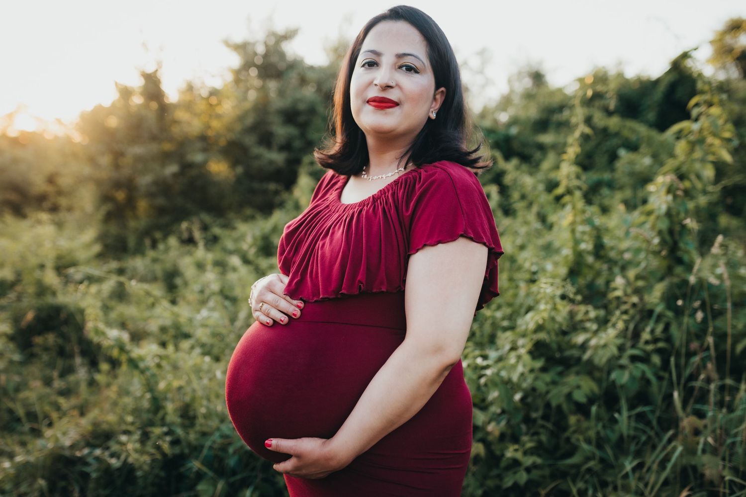 A pregnant person in a burgundy dress poses outdoors during golden hour surrounded by lush greenery.