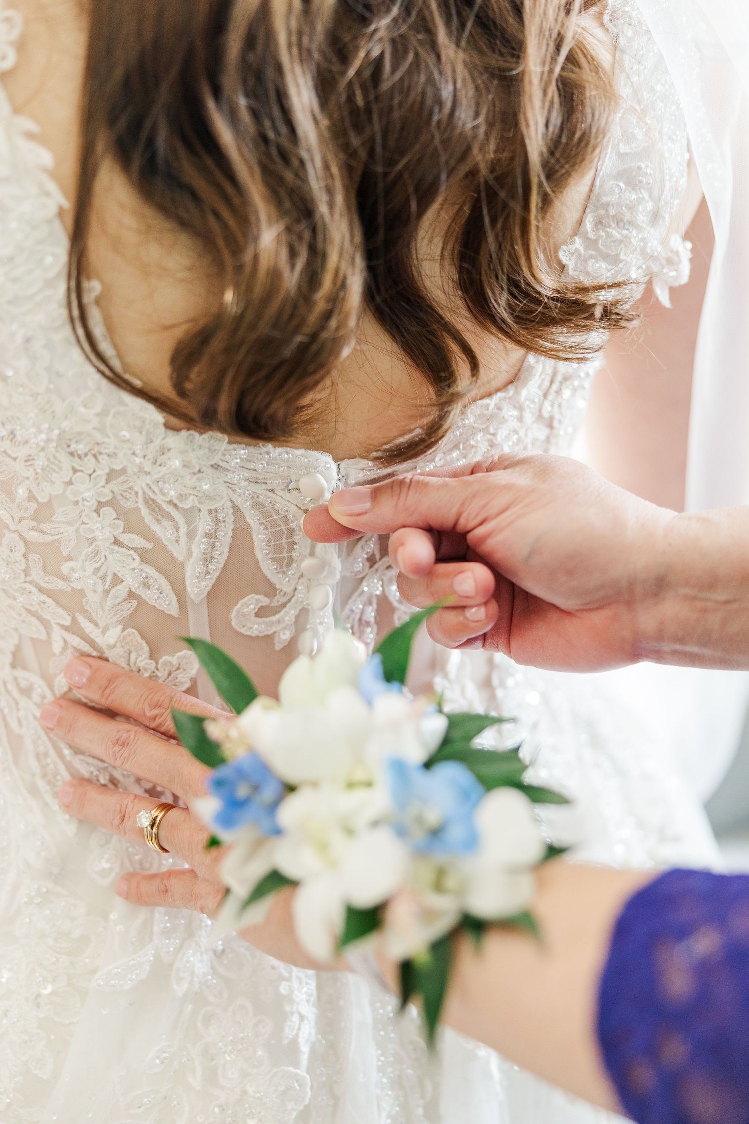 Someone helps button up an intricate lace wedding dress while holding a delicate white and blue floral bouquet.