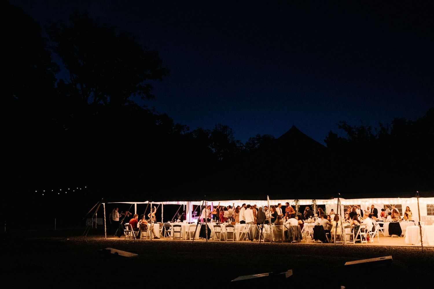 A glowing white tent illuminated at night hosts an outdoor wedding reception against a dark tree-lined sky.