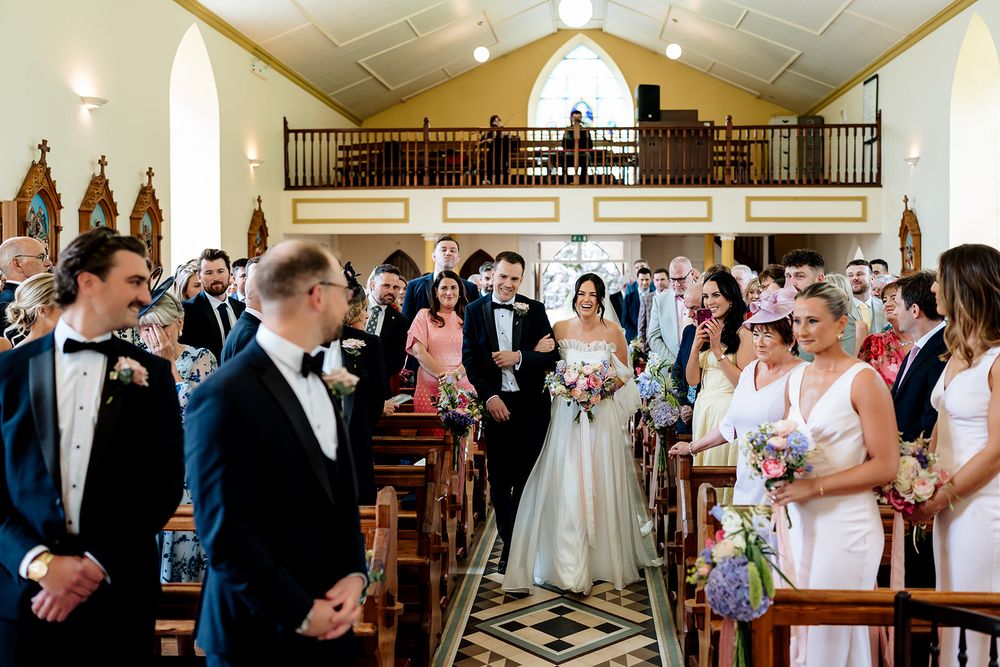 Wedding ceremony inside historic chapel with guests gathered around aisle as couple stands at altar with floral bouquets.