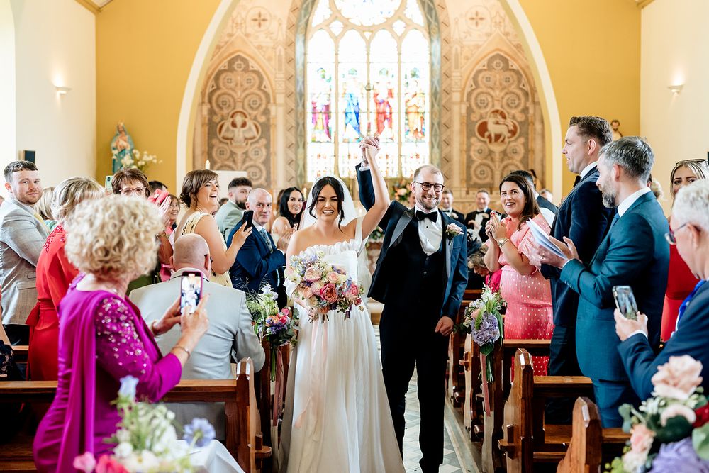A couple walks down the aisle at their wedding ceremony in a beautiful historic church as guests look on and celebrate.