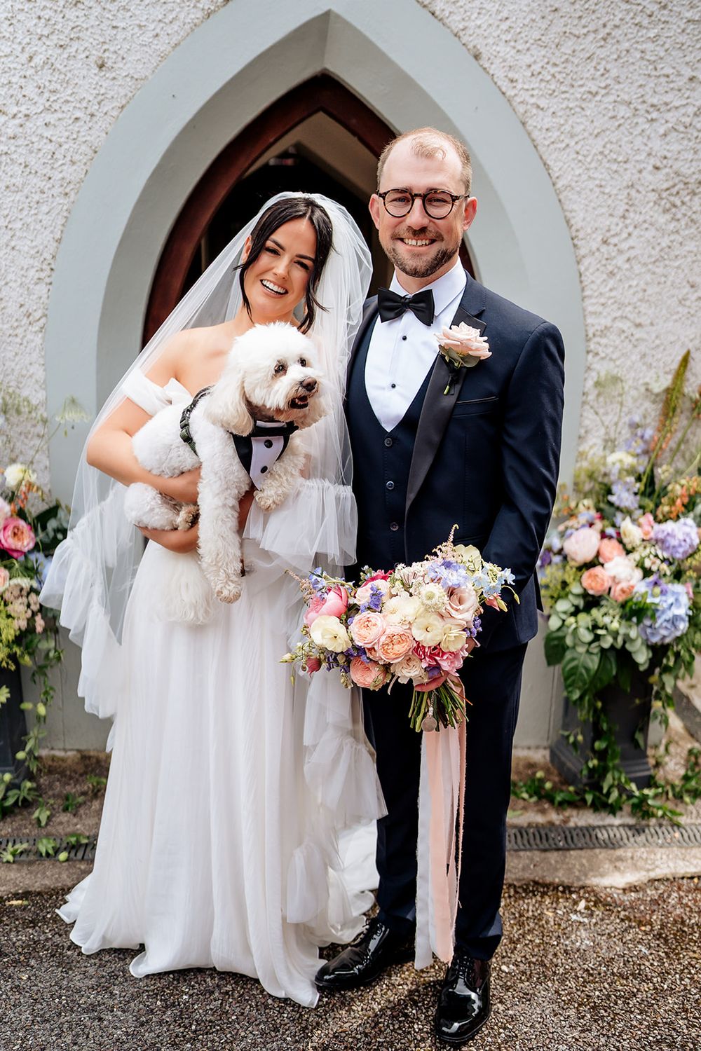 Happy newlyweds pose in front of church archway with their small white dog and pink floral bouquet.